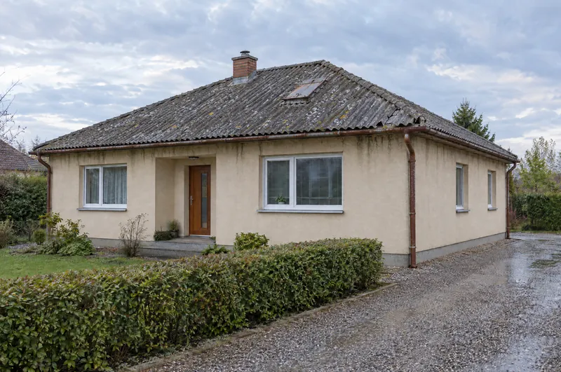 Přízemní dům s valbovou střechou ze starých šedých vlnitých desek s lišejníkem, prasklé rohy a rezavé žlaby.