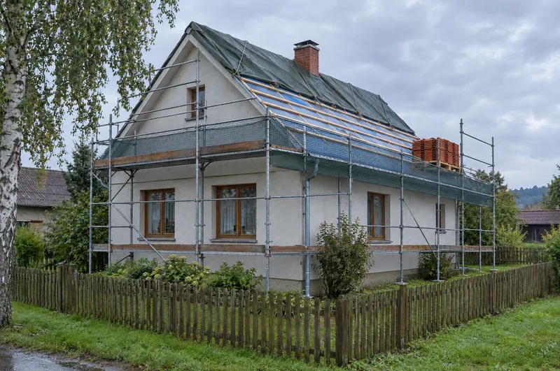 Tentýž dům jako v Křemenné během výměny střechy: lešení se sítí, polovina střechy na fólii a latích, horní část pod plachtou přivázanou lany, na lešení paleta pálených tašek.